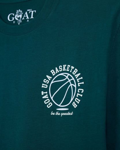 Close-up of a dark teal t-shirt featuring a white circular graphic on the chest with a basketball outline and the text 'GOAT USA BASKETBALL CLUB be the greatest'. The inside collar shows a white 'GOAT' logo.