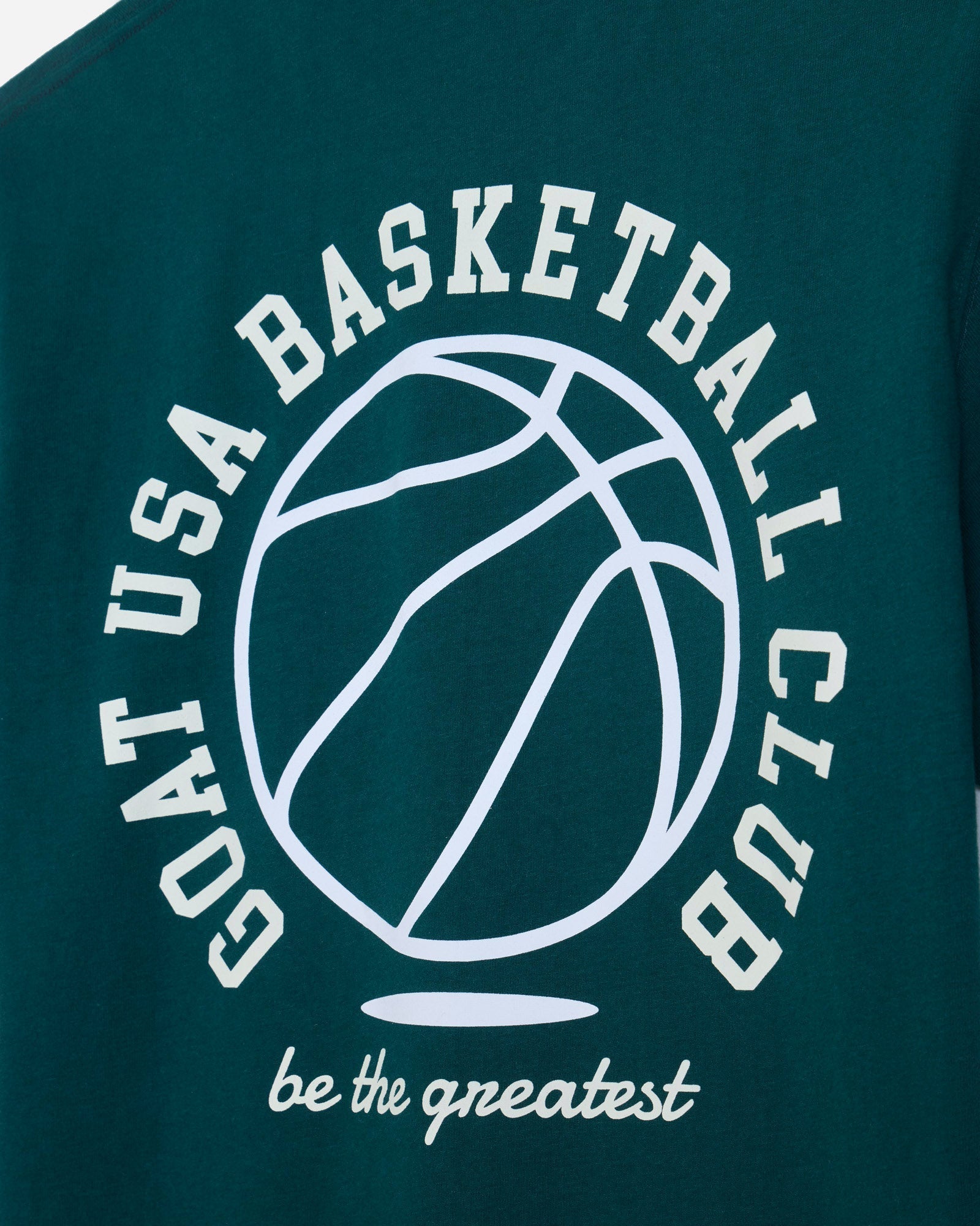 Close-up of a dark teal t-shirt featuring a white graphic print of a basketball and the text 'GOAT USA BASKETBALL CLUB' and 'be the greatest'.