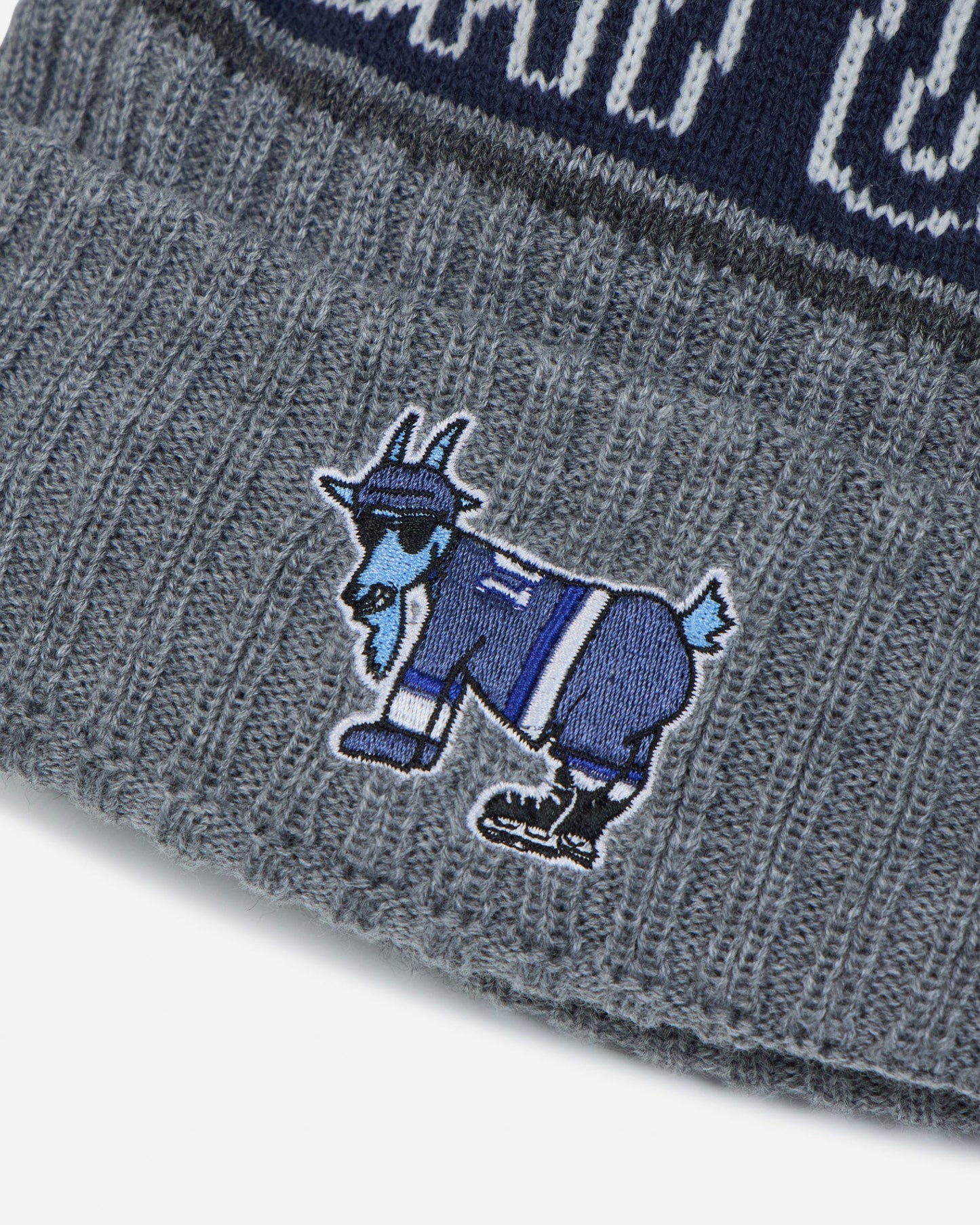 Close-up of a gray knit beanie with a blue embroidered goat mascot logo on the cuff, showing part of a navy and gray patterned band at the top.