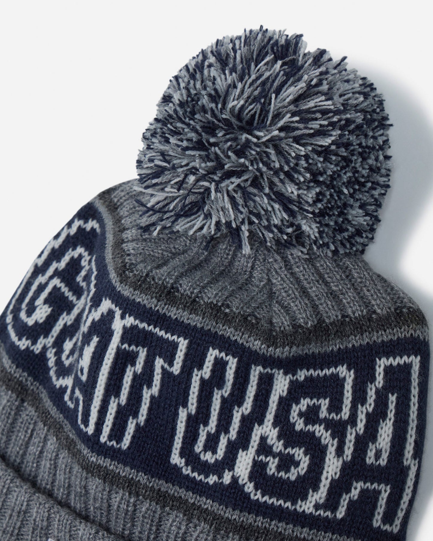 A close-up of a knit pom-pom beanie in heather gray and navy blue, featuring the word 'GOAT USA' knitted across the front in white.