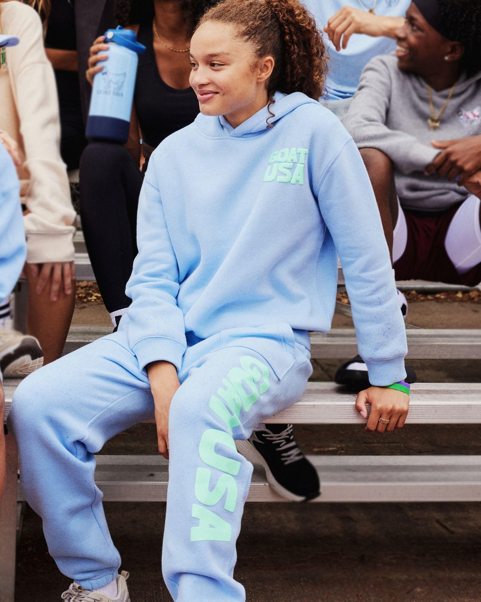 Person wearing a light blue tracksuit with 'GOAT USA' text, sitting on steps.#color_carolina-blue