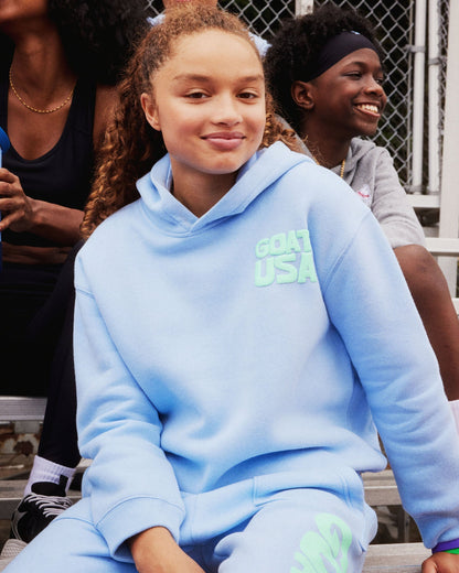 Person wearing a light blue hoodie with 'GOAT USA' text, sitting outdoors with others.#color_carolina-blue