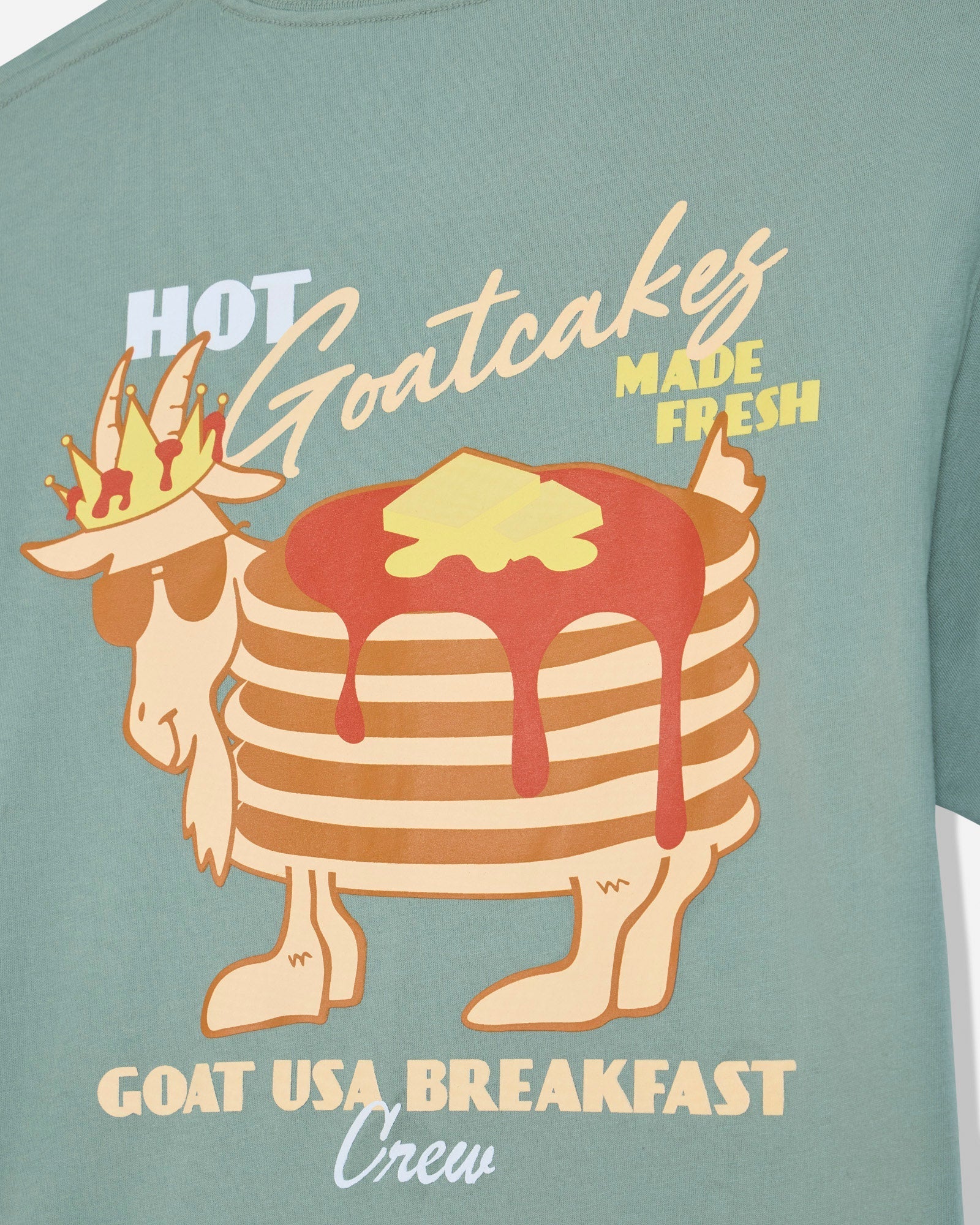 A close-up of a sage green t-shirt featuring a graphic of a goat wearing sunglasses and a crown, with a stack of pancakes topped with syrup and butter. The text reads 'HOT Goatcakes MADE FRESH' and 'GOAT USA BREAKFAST Crew'.