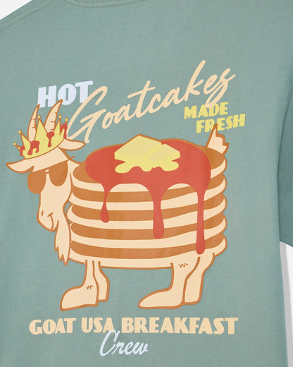 A close-up of a sage green t-shirt featuring a graphic of a goat wearing sunglasses and a crown, with a stack of pancakes topped with syrup and butter. The text reads 'HOT Goatcakes MADE FRESH' and 'GOAT USA BREAKFAST Crew'.