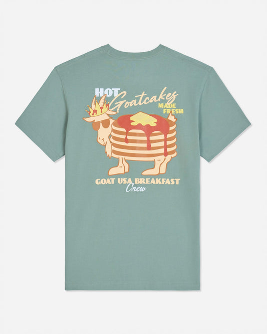 A sage green short-sleeve t-shirt with a retro graphic of a stack of pancakes wearing a crown and sunglasses, with the text 'HOT Goatcakes MADE FRESH' and 'GOAT USA BREAKFAST Union' on a light gray background.