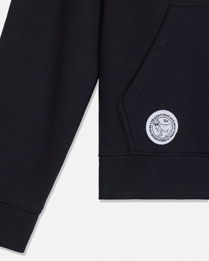 Close-up of a black crewneck sweatshirt showing the left sleeve and chest area, featuring a circular white embroidered logo patch on the pocket.