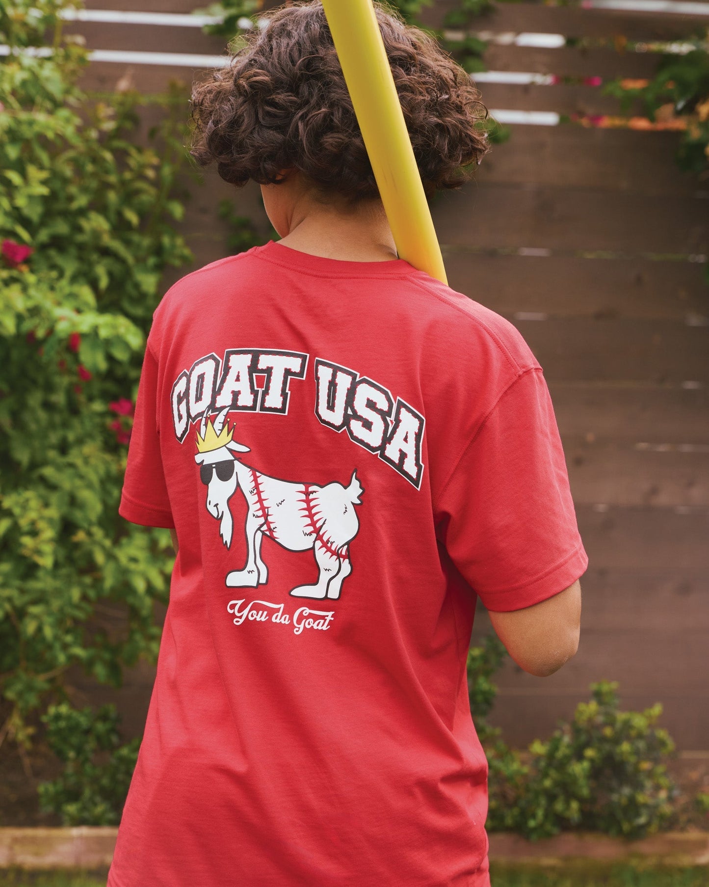Person wearing a red 'GOAT USA' t-shirt with a graphic design, standing outdoors.