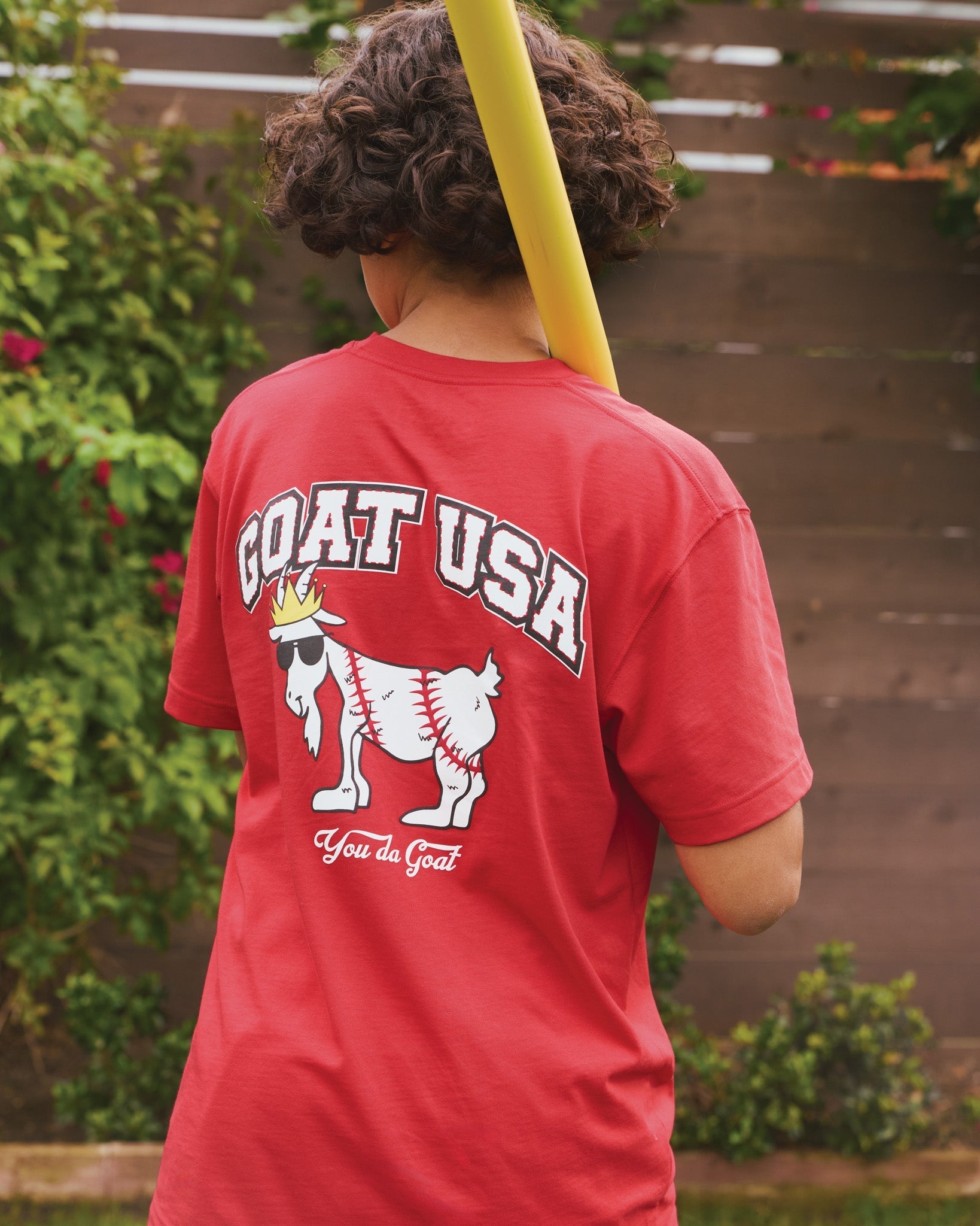 Person wearing a red 'GOAT USA' t-shirt with a graphic design, standing outdoors.
