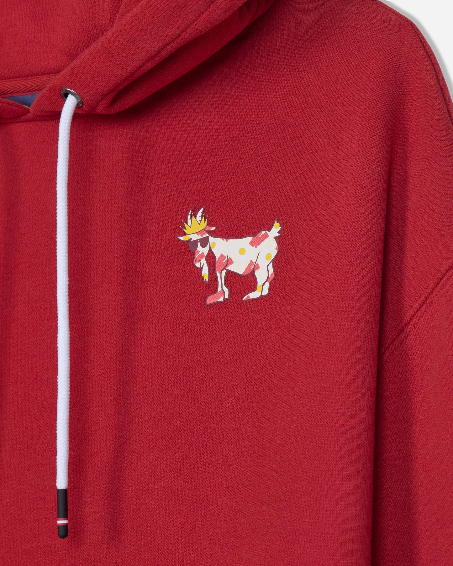 A close-up of a red hooded sweatshirt featuring a white drawstring and a small colorful embroidered goat character on the left chest.