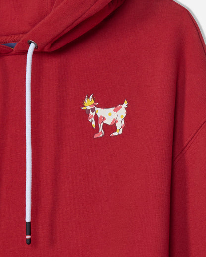 A close-up of a red hooded sweatshirt featuring a white drawstring and a small colorful embroidered goat character on the left chest.