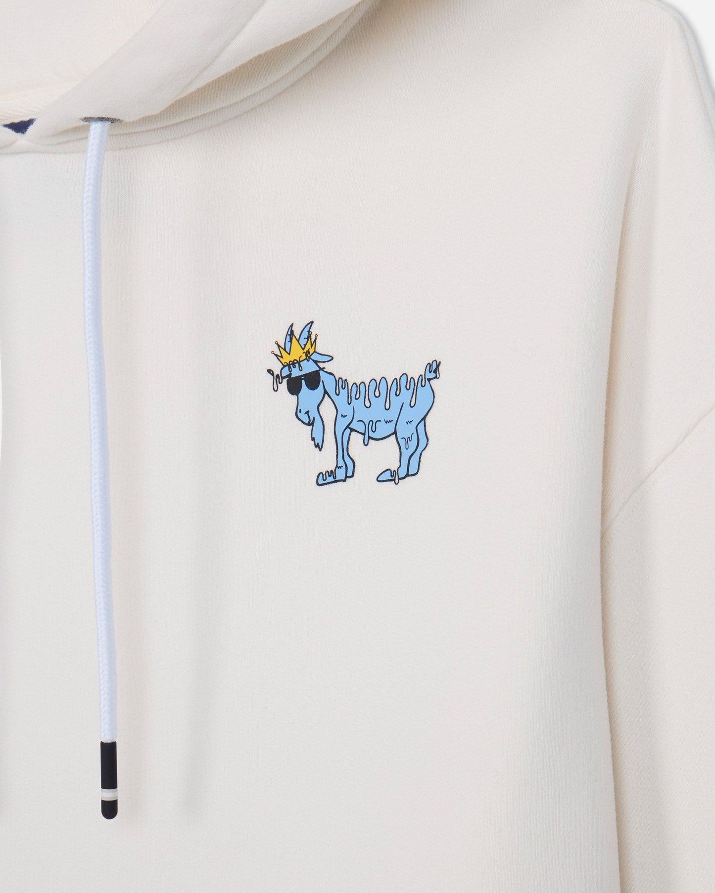 Close-up of a cream-colored hoodie featuring a small graphic of a blue goat wearing sunglasses and a gold crown on the left chest, with a white drawstring visible.
