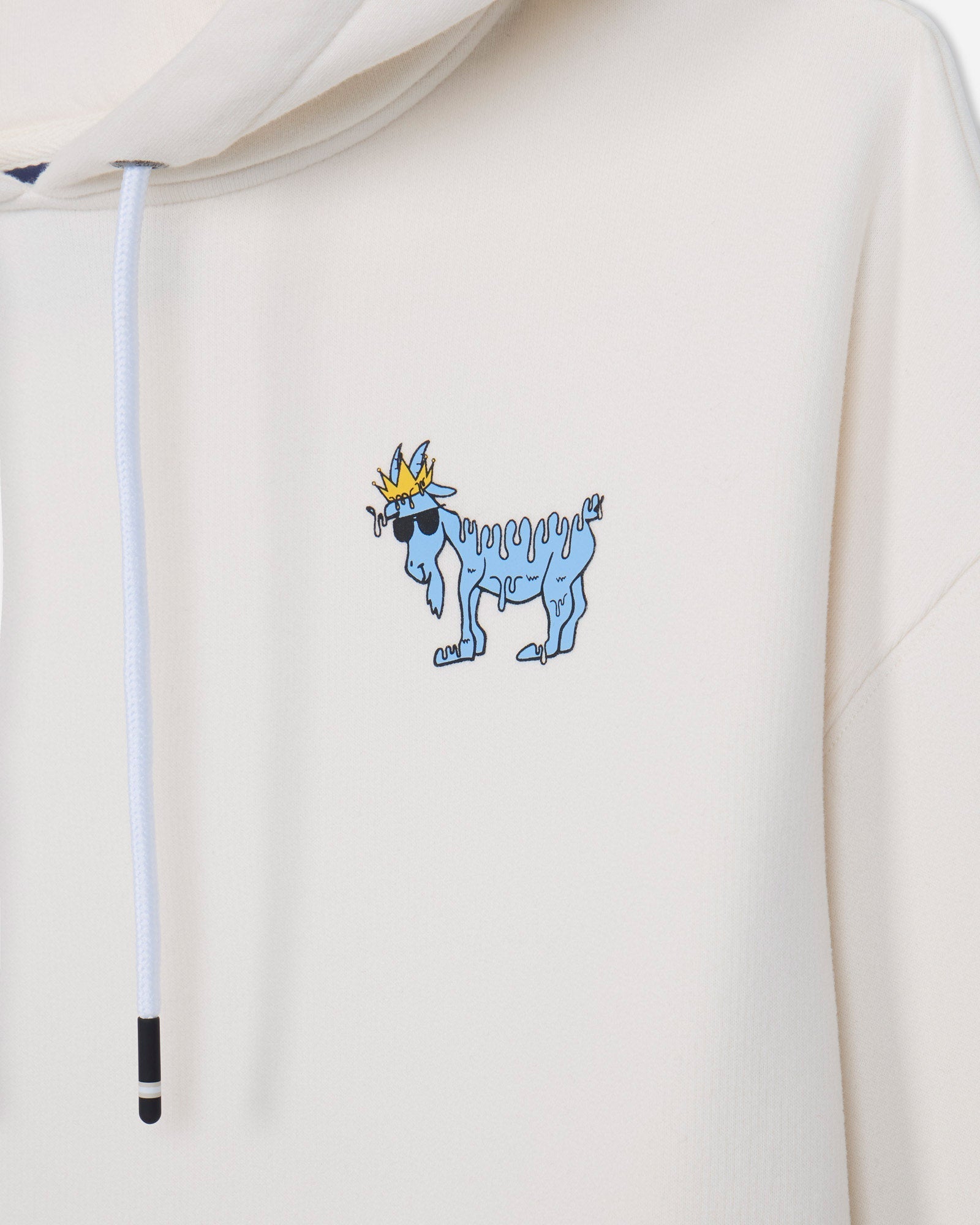 Close-up of a cream-colored hoodie featuring a small graphic of a blue goat wearing sunglasses and a gold crown on the left chest, with a white drawstring visible.