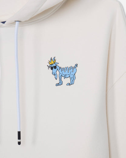 Close-up of a cream-colored hoodie featuring a small graphic of a blue goat wearing sunglasses and a gold crown on the left chest, with a white drawstring visible.