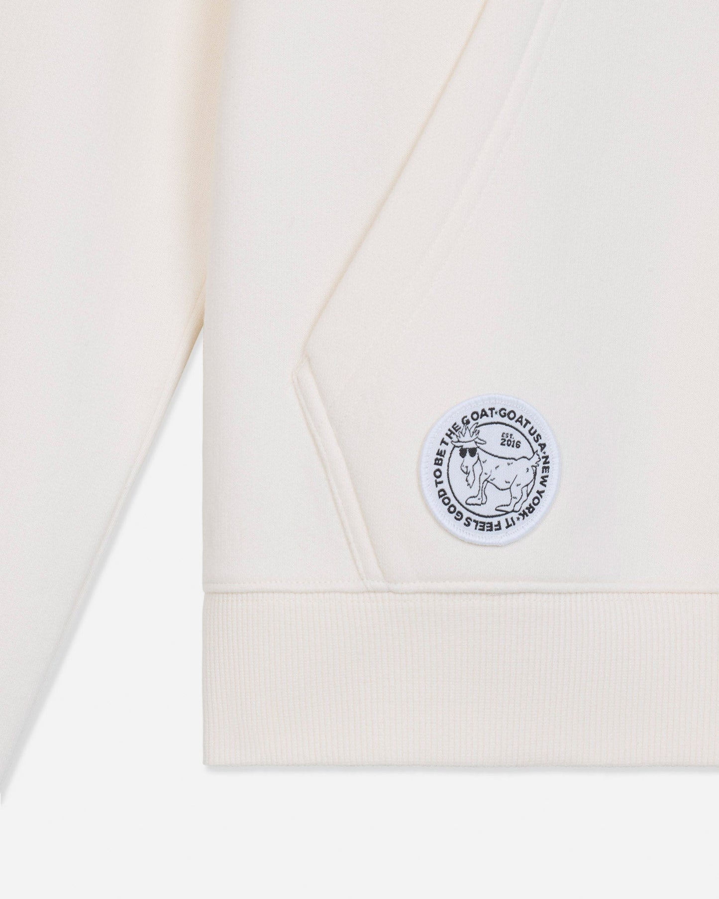 Close-up of a cream-colored sweatshirt showing the sleeve cuff, ribbed hem, and a circular embroidered patch on the chest.