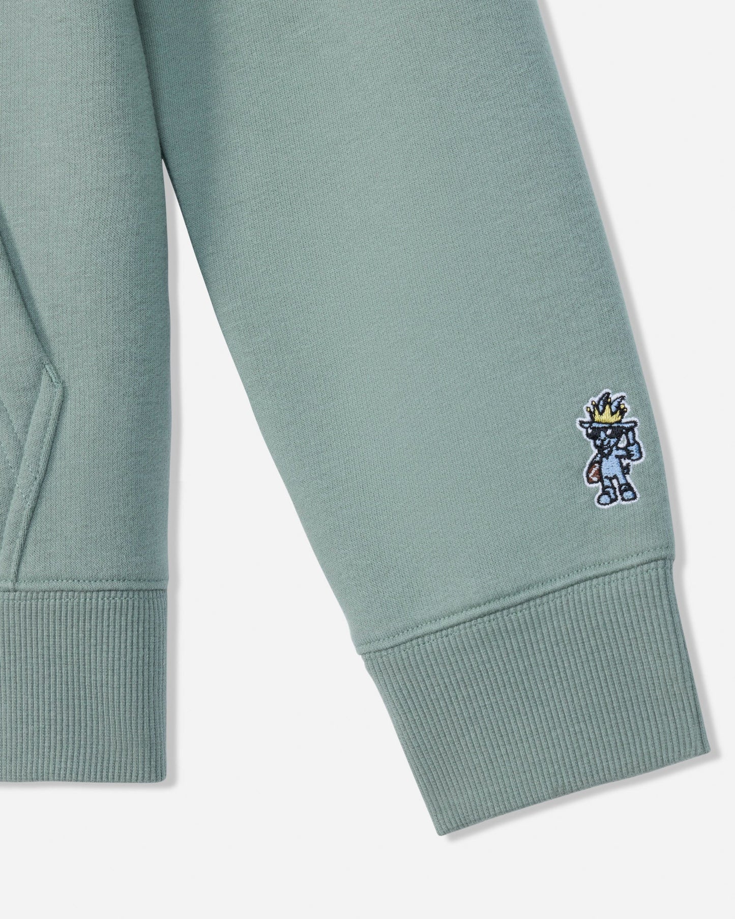 Close-up of a sage green sweatshirt sleeve with a ribbed cuff, featuring a small embroidered cartoon character on the cuff.