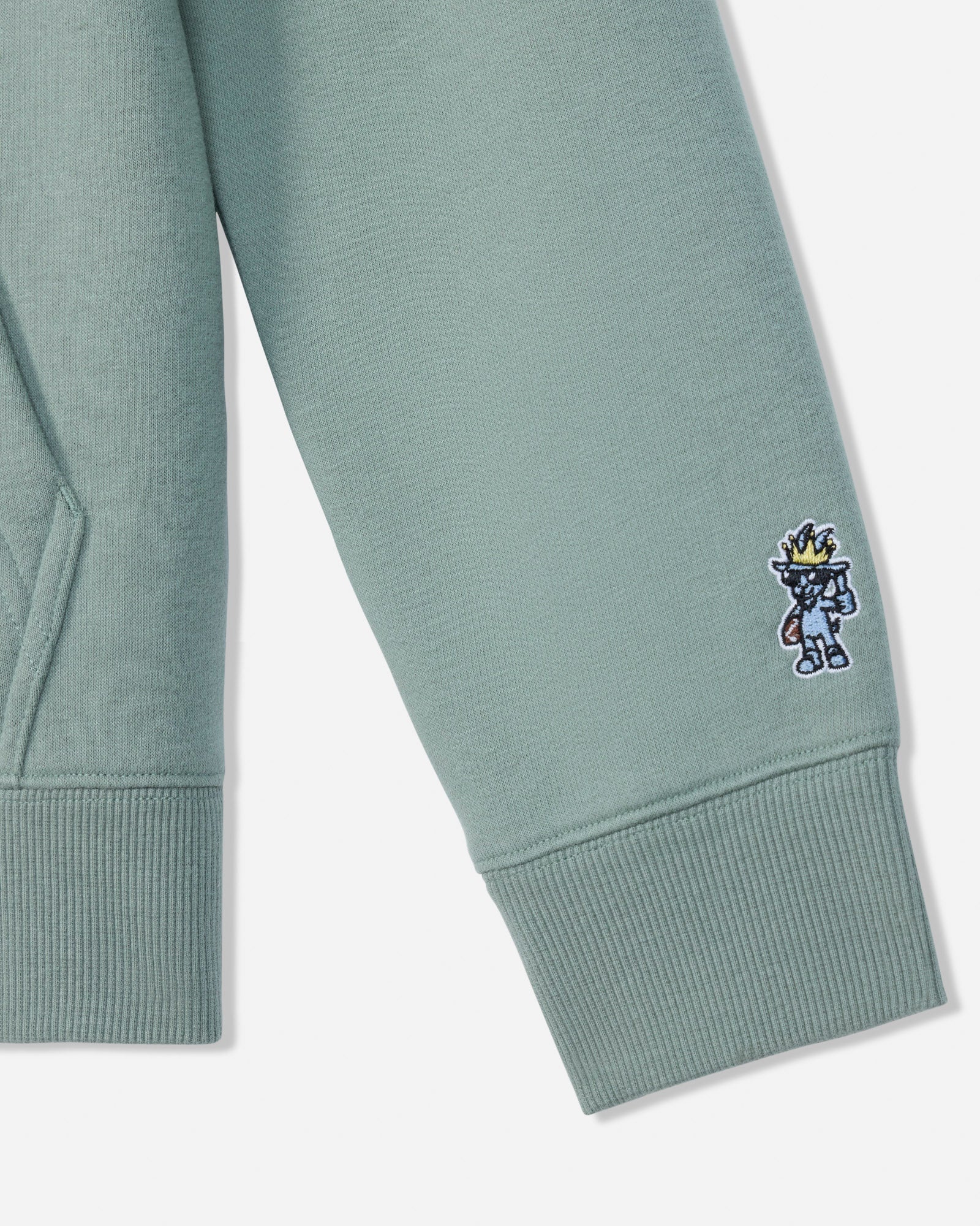 Close-up of a sage green sweatshirt sleeve with a ribbed cuff, featuring a small embroidered cartoon character on the cuff.
