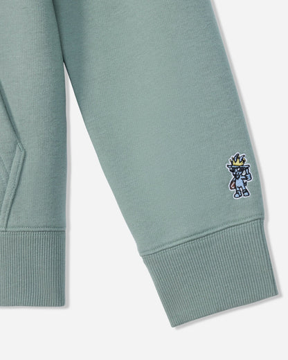 Close-up of a sage green sweatshirt sleeve with a ribbed cuff, featuring a small embroidered cartoon character on the cuff.