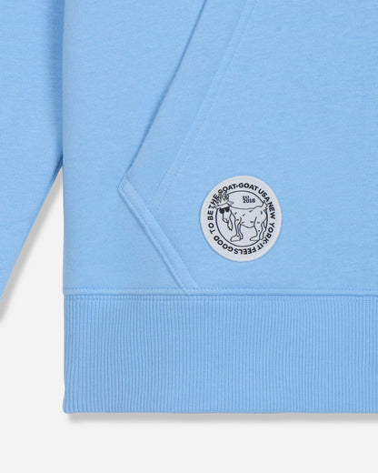 Close-up of a light blue sweatshirt showing the sleeve, ribbed cuff, and a circular white patch with black text and a cartoon character on the pocket.