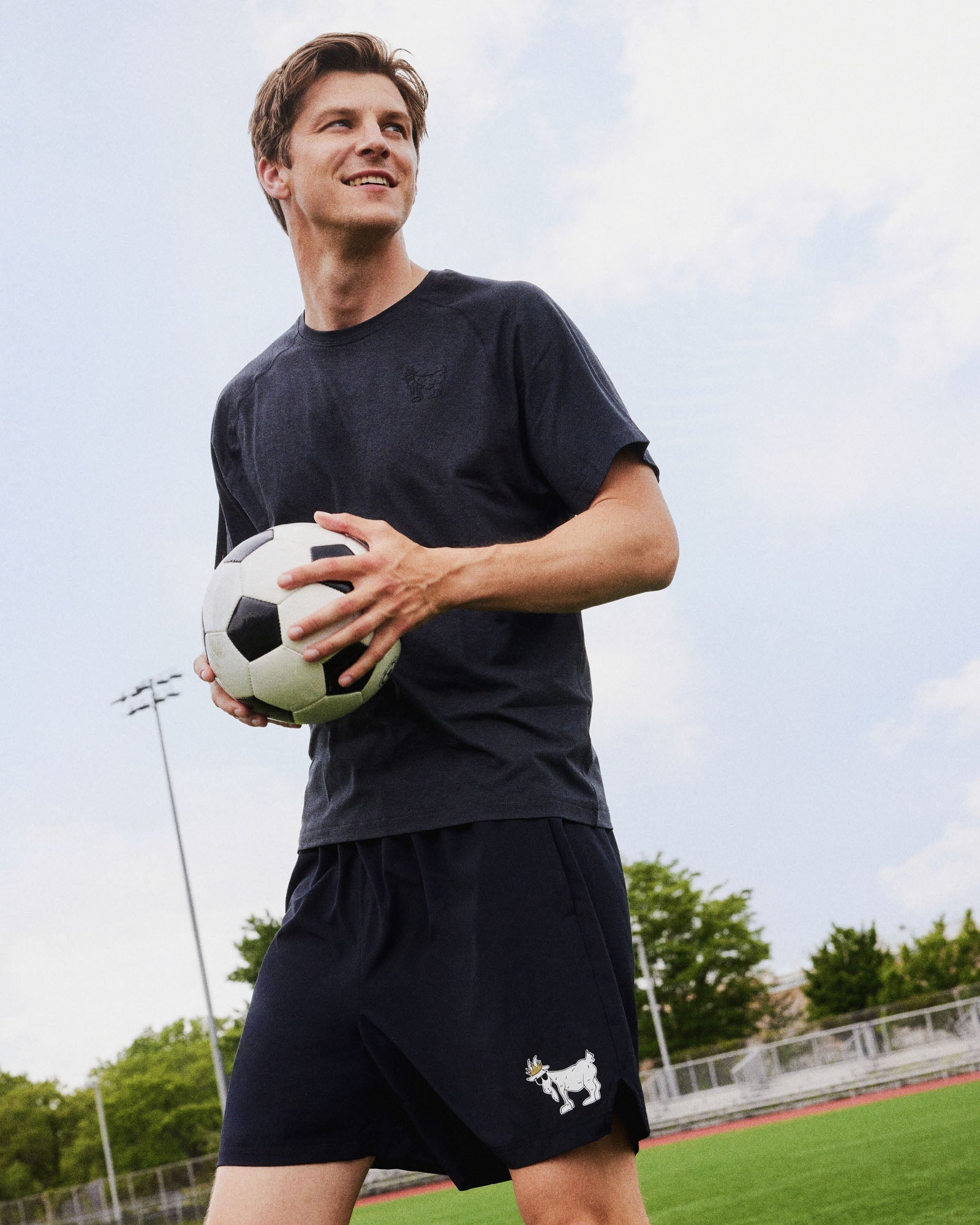Man holding a soccer ball on a sports field with trees and sky in the background#color_black