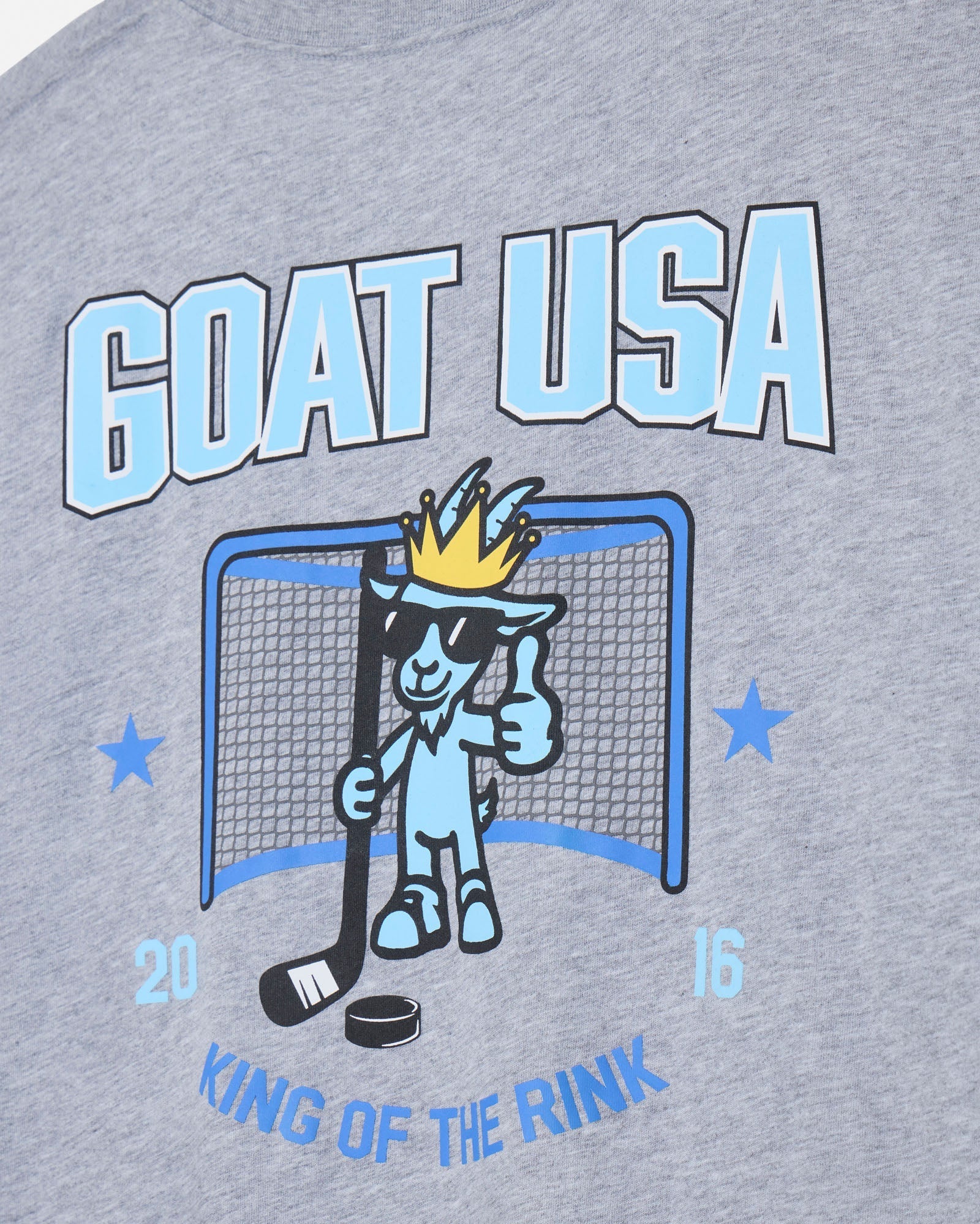 Close-up of a heather gray graphic T-shirt featuring a light blue and black 'GOAT USA' design with a cartoon goat in a sunglasses and crown, plus 'KING OF THE RINK' text.
