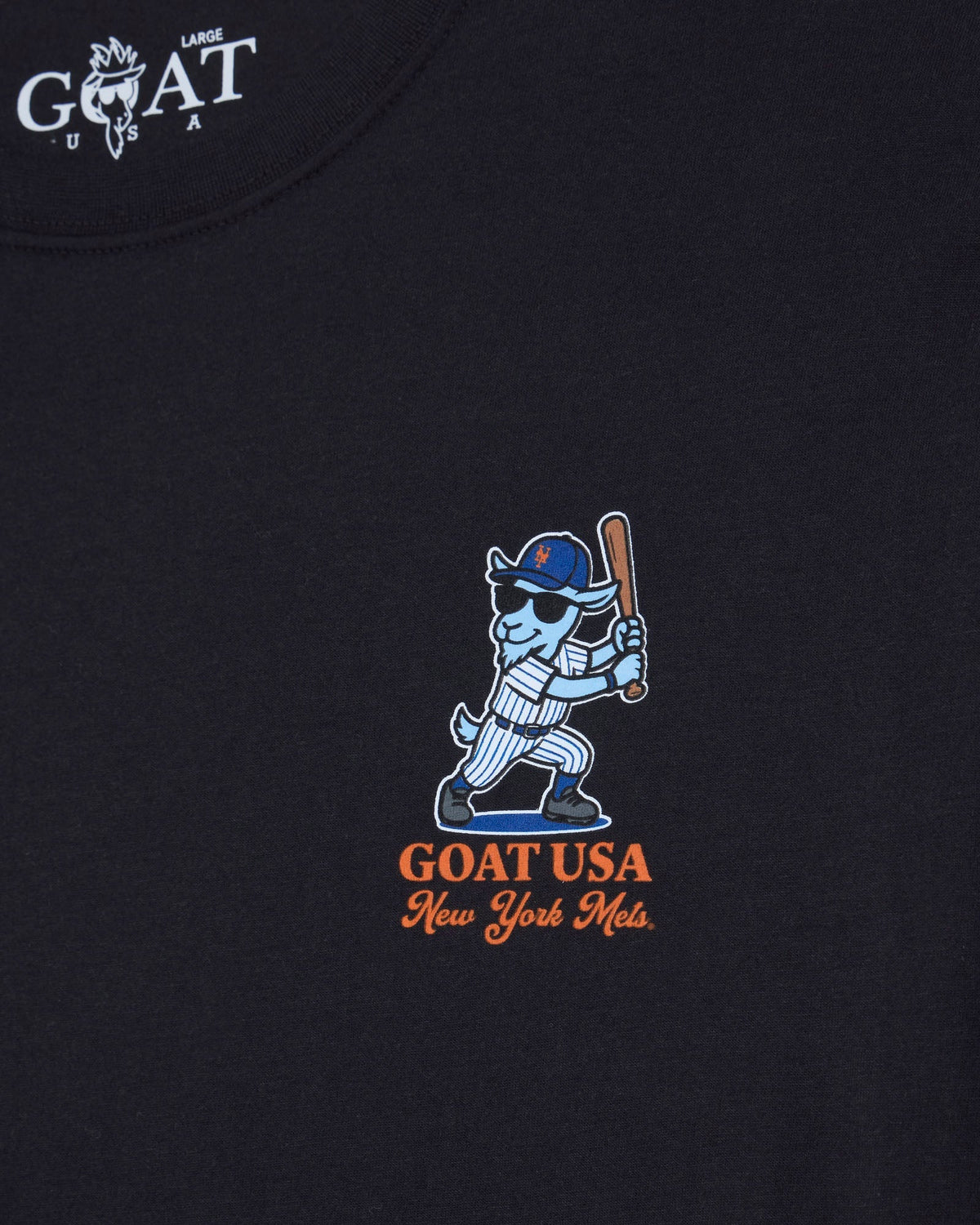 Close-up of a black t-shirt featuring a central graphic of a blue and white goat mascot holding a baseball bat, with 'GOAT USA New York Mets' below in orange. The top left corner shows a white 'GOAT USA LARGE' brand print.