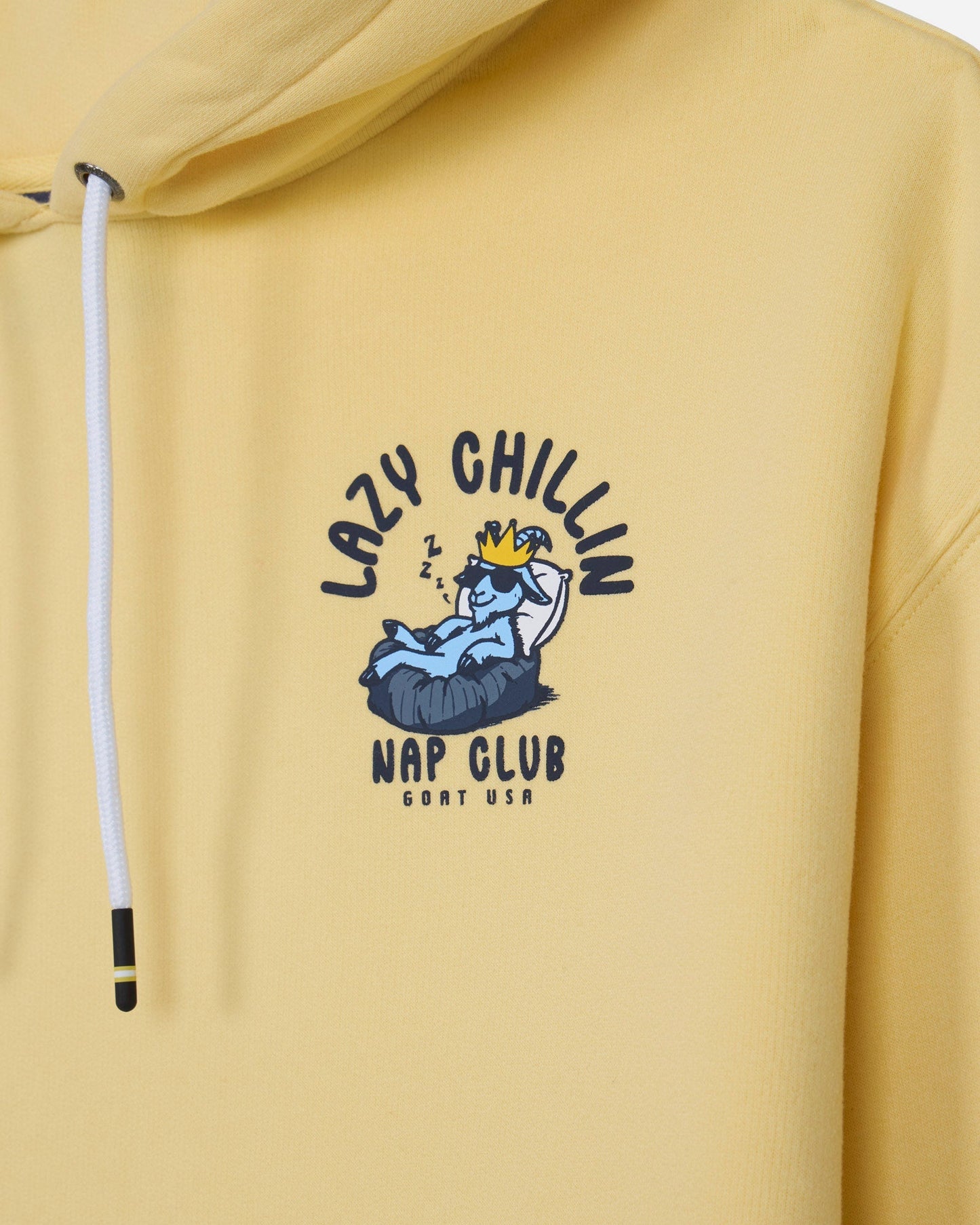 A close-up of a pale yellow hoodie featuring a central graphic print of a cartoon blue character wearing a gold crown, with the text 'LAZY CHILLIN' and 'NAP CLUB' in dark blue.
