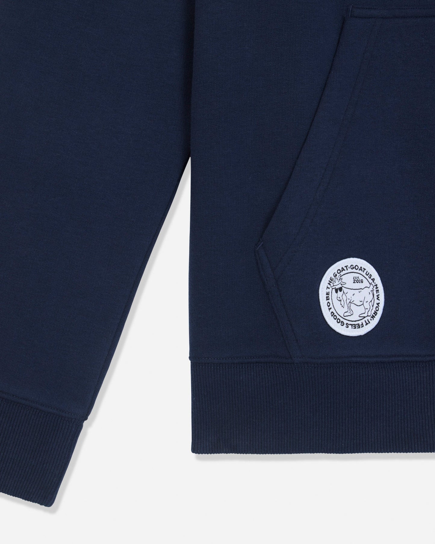 Close-up of a navy blue sweatshirt showing the sleeve, ribbed hem, and a circular white embroidered logo patch on the lower front.#color_navy-blazer