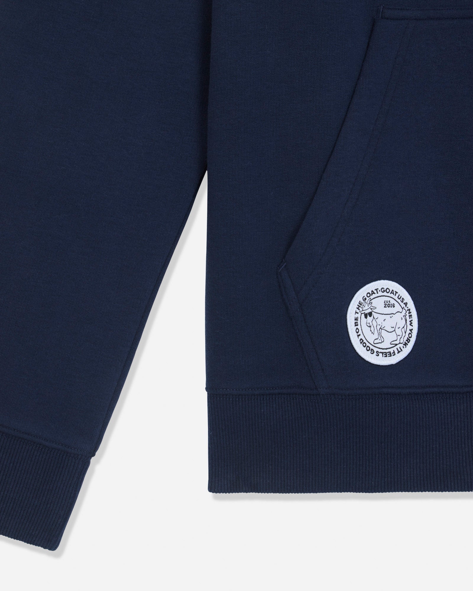 Close-up of a navy blue sweatshirt showing the sleeve, ribbed hem, and a circular white embroidered logo patch on the lower front.#color_navy-blazer