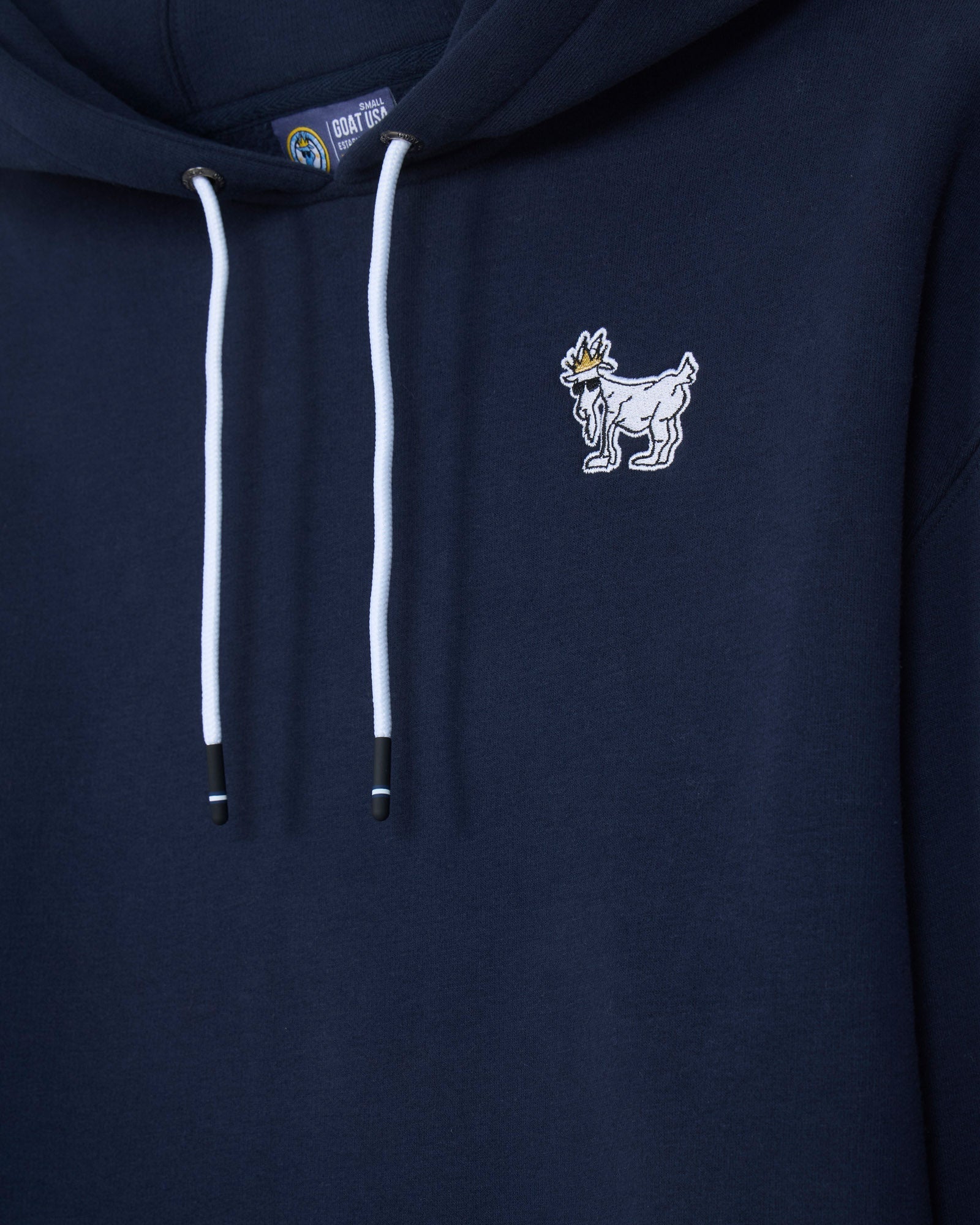 Close-up of a navy blue hoodie with white drawstrings and a small white embroidered goat mascot logo on the left chest.#color_navy-blazer