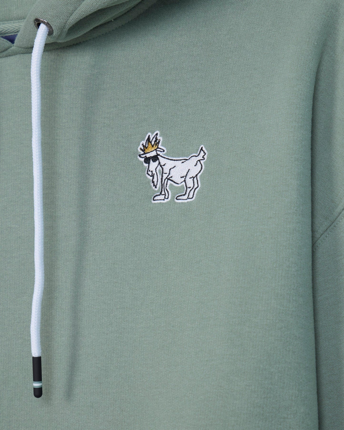 Close-up of a sage green hoodie featuring a small embroidered goat logo with a crown on the chest and a white drawstring.#color_steeped-sage