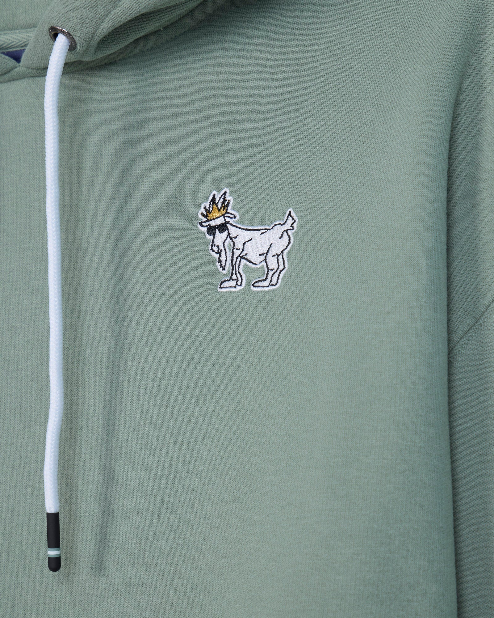 Close-up of a sage green hoodie featuring a small embroidered goat logo with a crown on the chest and a white drawstring.#color_steeped-sage