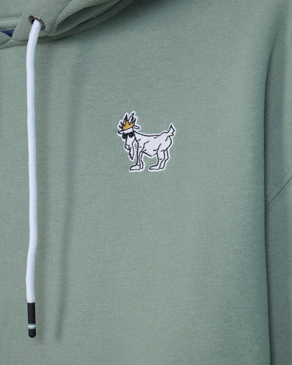 Close-up of a sage green hoodie featuring a small embroidered goat logo with a crown on the chest and a white drawstring.#color_steeped-sage