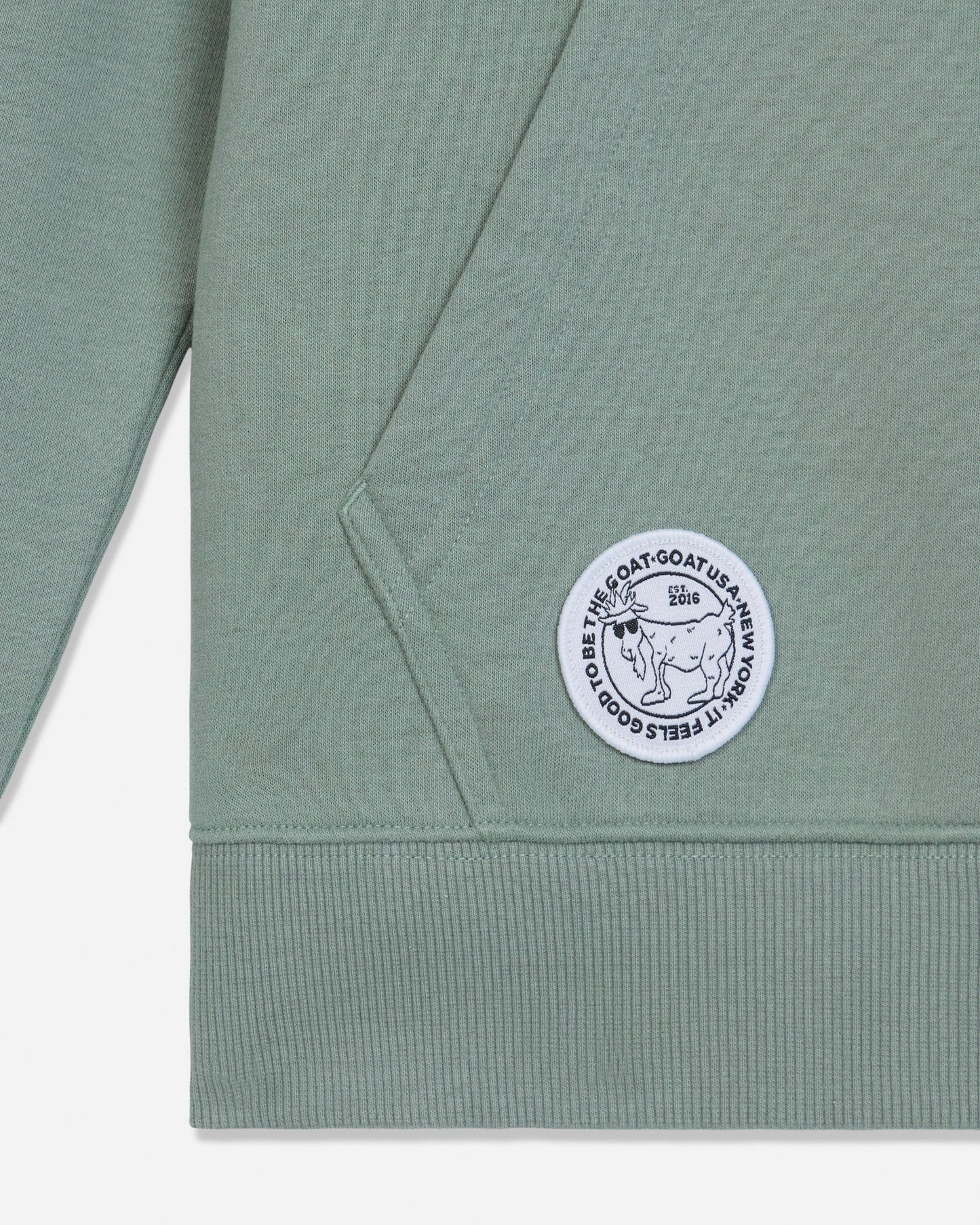 Close-up of a sage green sweatshirt showing the sleeve, ribbed cuff, and a circular white embroidered patch with a goat illustration and text.#color_steeped-sage