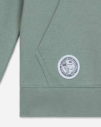 Close-up of a sage green sweatshirt showing the sleeve, ribbed cuff, and a circular white embroidered patch with a goat illustration and text.#color_steeped-sage