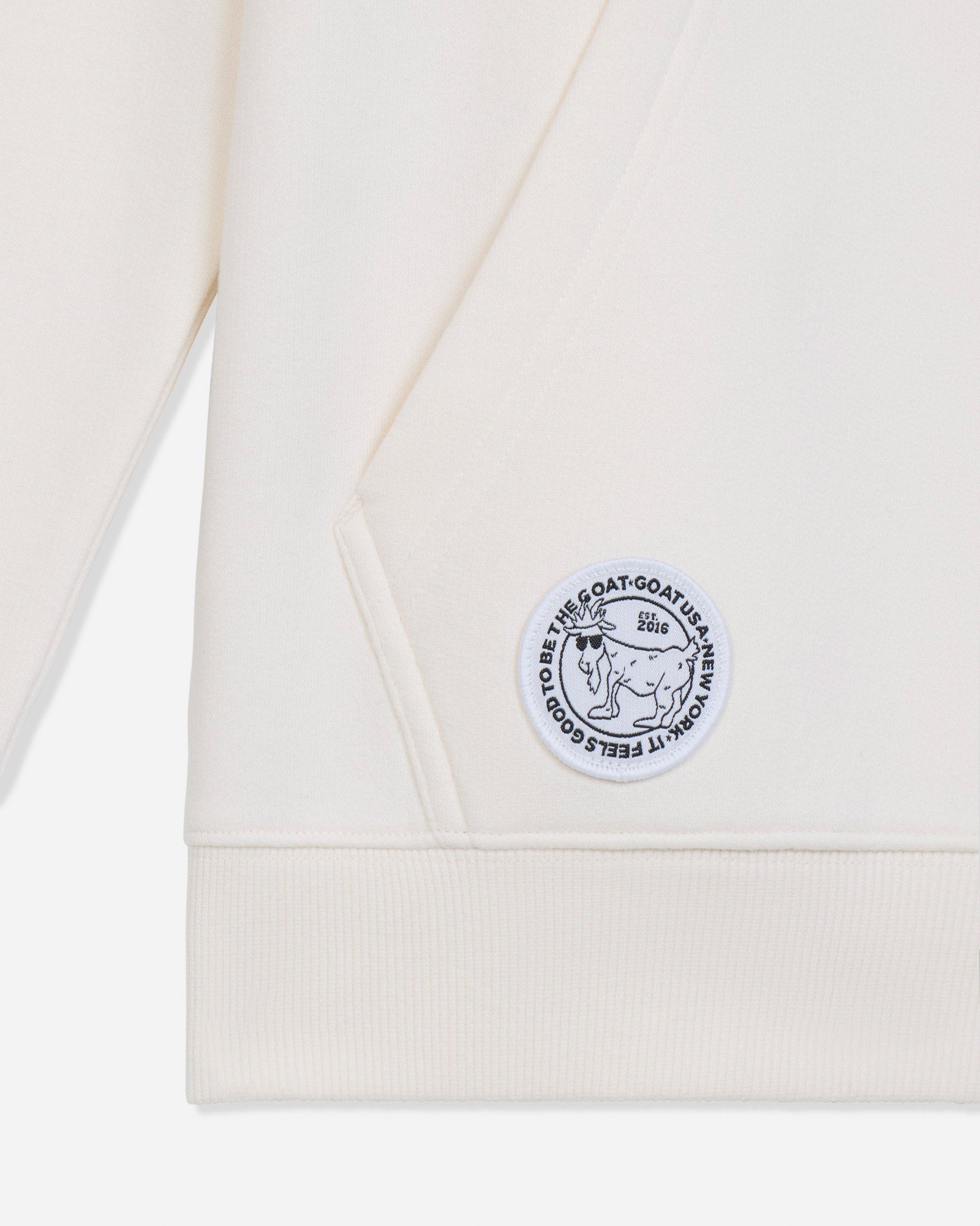 Close-up of an off-white ribbed knit garment, likely a sweatshirt or hoodie, showing a circular embroidered patch with a goat illustration and text.#color_bone