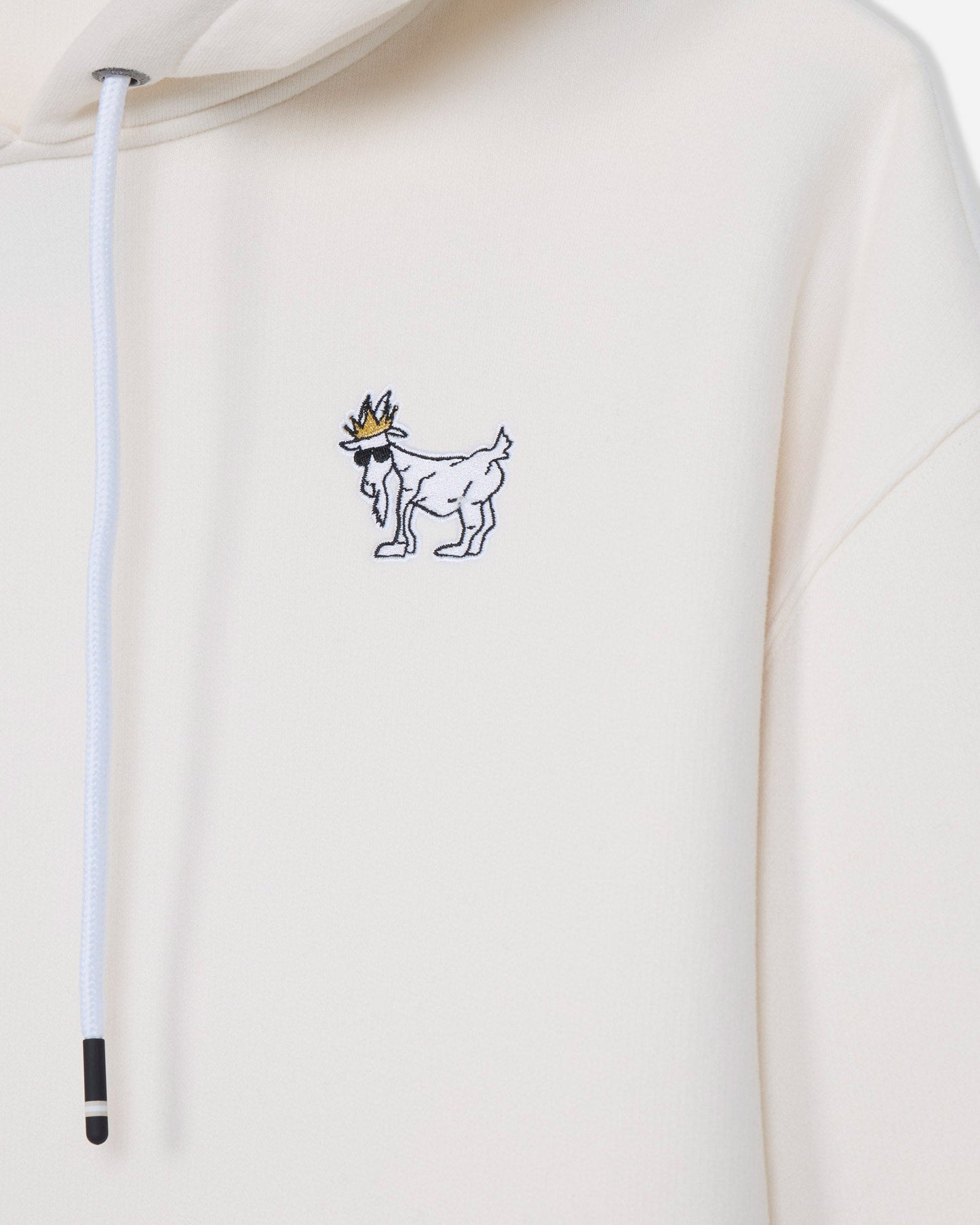 Close-up of a cream-colored hoodie featuring a small embroidered graphic of a white goat wearing a gold crown on the left chest, with a white drawstring hanging on the left side.#color_bone