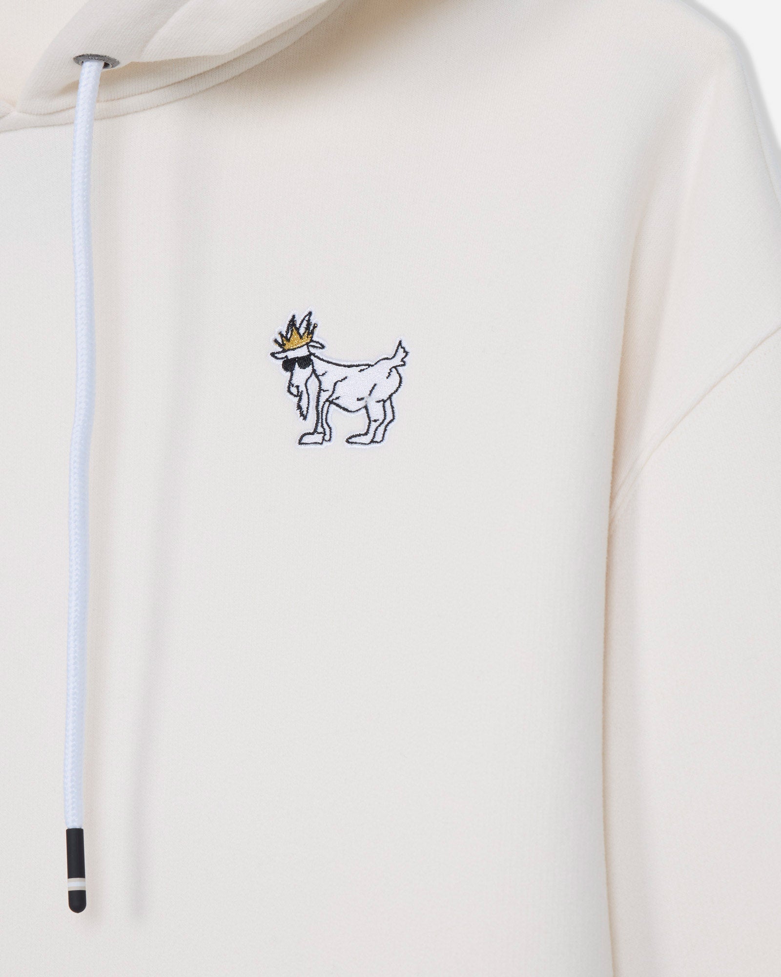 Close-up of a cream-colored hoodie featuring a small embroidered graphic of a white goat wearing a gold crown on the left chest, with a white drawstring hanging on the left side.#color_bone