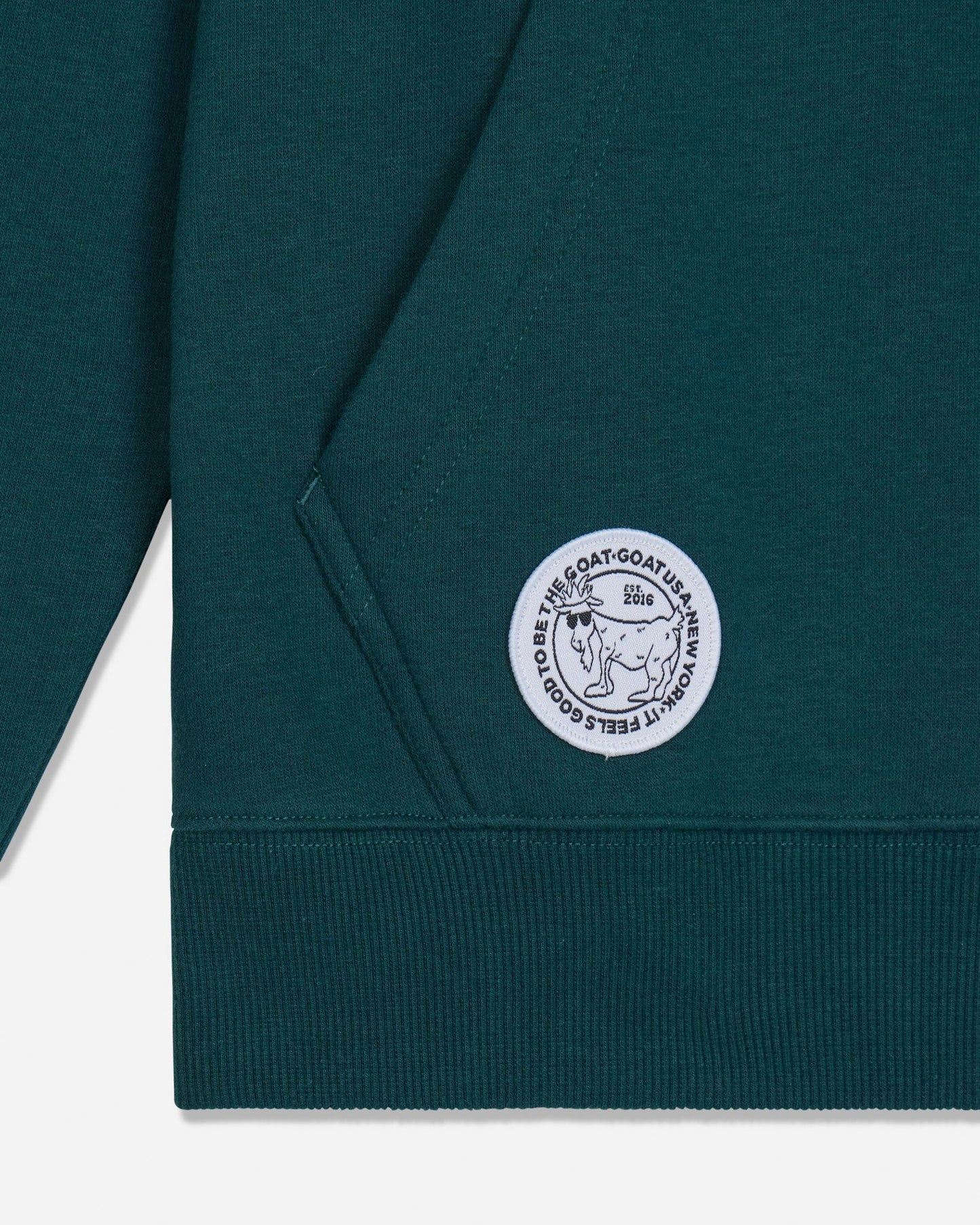 Close-up of a dark teal-green sweatshirt showing the sleeve, ribbed cuff, and a circular white embroidered patch on the pocket with the text 'IT FEELS GOOD TO BE THE GOAT' and a goat illustration.