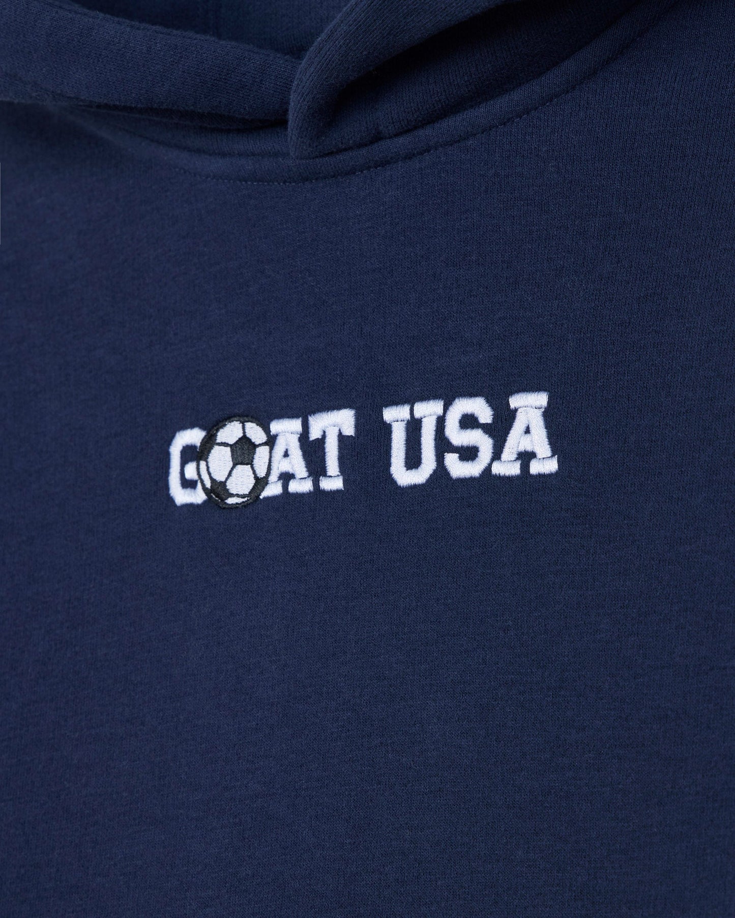 Close-up of a navy blue hoodie with 'GOAT USA' embroidered in white across the chest, where the letter O is replaced by a black-and-white soccer ball graphic.