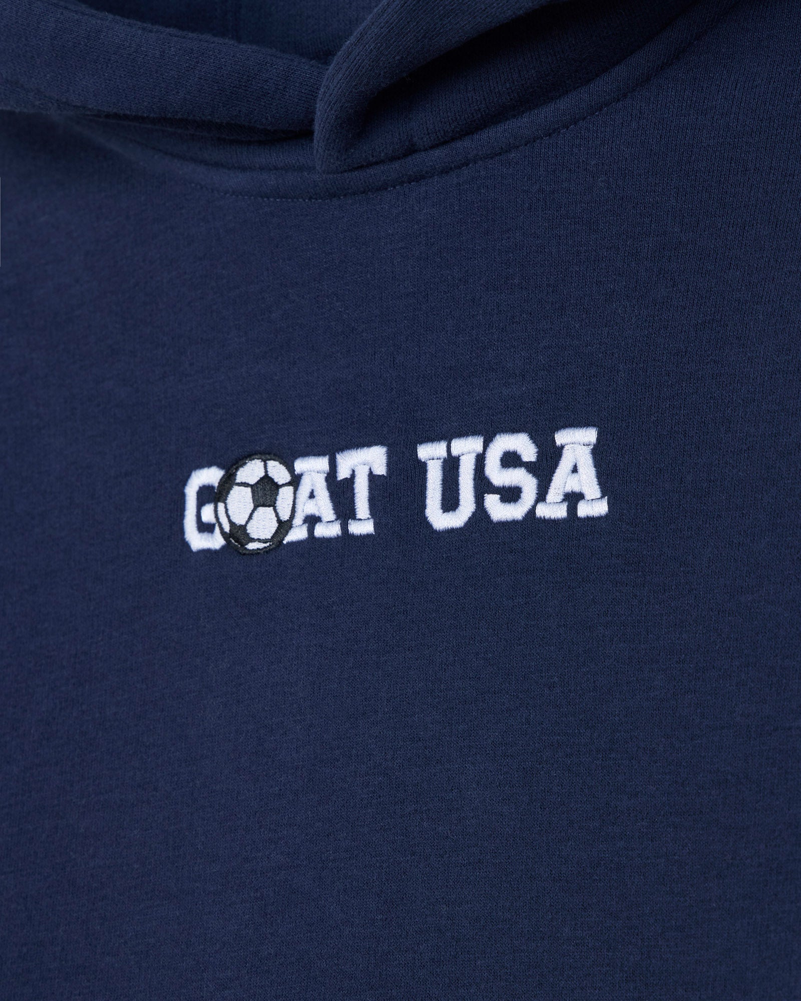 Close-up of a navy blue hoodie with 'GOAT USA' embroidered in white across the chest, where the letter O is replaced by a black-and-white soccer ball graphic.