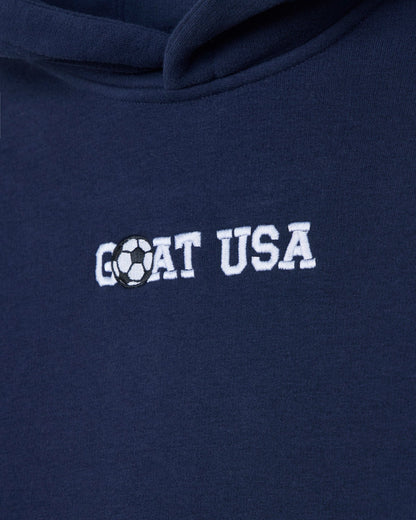 Close-up of a navy blue hoodie with 'GOAT USA' embroidered in white across the chest, where the letter O is replaced by a black-and-white soccer ball graphic.