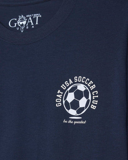Close-up of a navy blue t-shirt featuring a white printed graphic of a soccer ball with the text 'GOAT USA SOCCER CLUB' and 'be the greatest' below it.