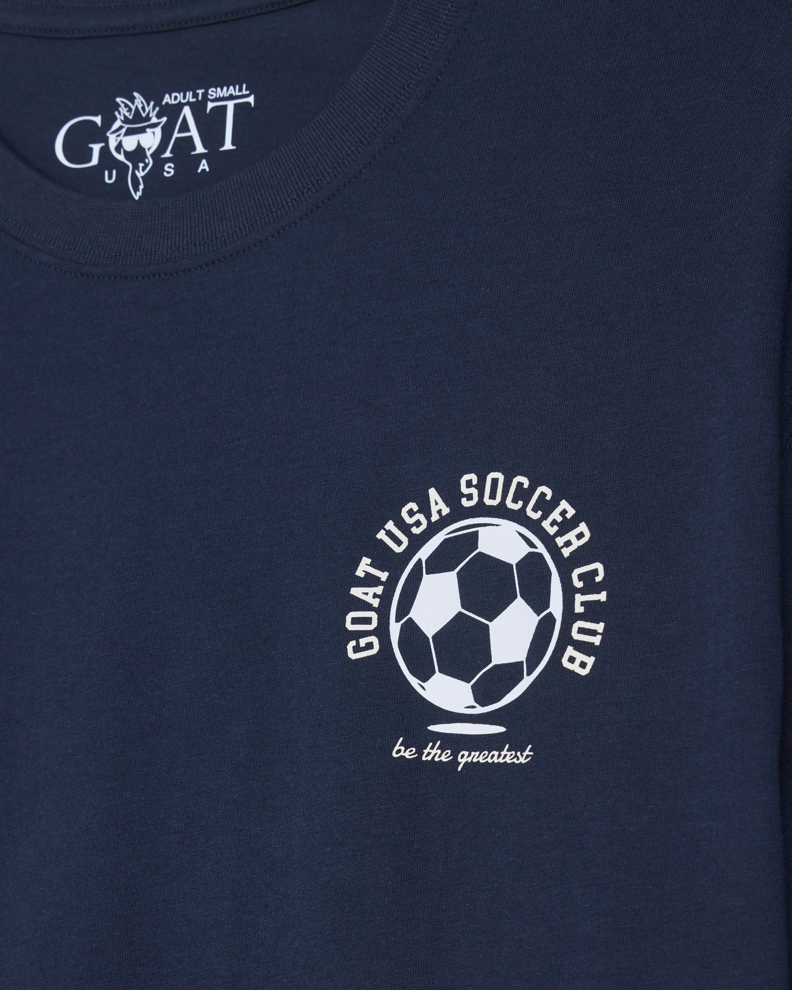 Close-up of a navy blue t-shirt featuring a white printed graphic of a soccer ball with the text 'GOAT USA SOCCER CLUB' and 'be the greatest' below it.