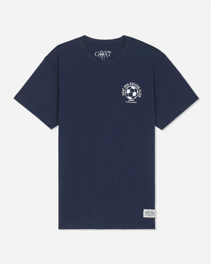 A navy blue short-sleeve t-shirt with a white circular soccer-themed graphic on the left chest and a white 'GOAT' brand print inside the collar, displayed on a light gray background.