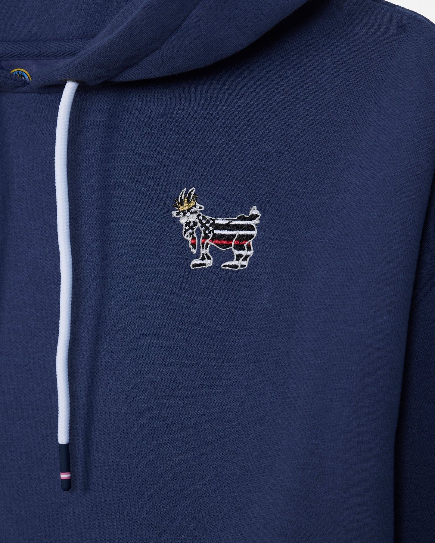 Close-up of a navy blue hoodie featuring a white drawstring and a small embroidered goat graphic on the chest with red, white, and black stripes and a gold crown.