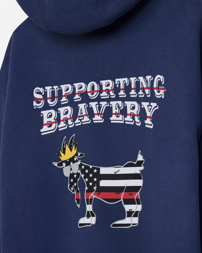 Close-up of the back of a navy blue hoodie featuring a graphic design with the words 'SUPPORTING BRAVERY' in white outlined letters with red strike-through lines, and a goat illustration filled with an American flag pattern and a gold crown.