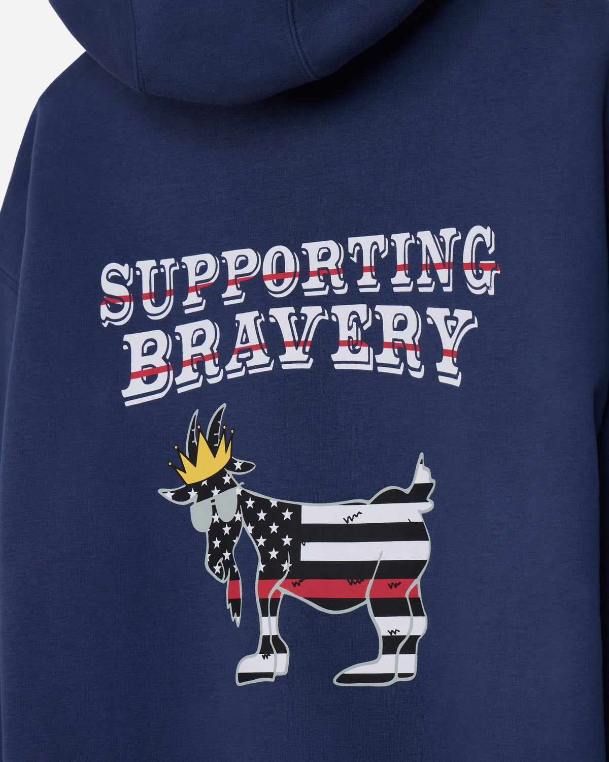 Close-up of the back of a navy blue hoodie featuring a graphic design with the words 'SUPPORTING BRAVERY' in white outlined letters with red strike-through lines, and a goat illustration filled with an American flag pattern and a gold crown.