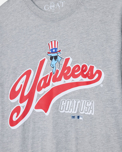 Close-up of a heather gray t-shirt featuring a large red and white 'Yankees' script logo with a blue cartoon character wearing a red, white, and blue top hat. The text 'GOAT USA' is printed below in white with a blue outline.