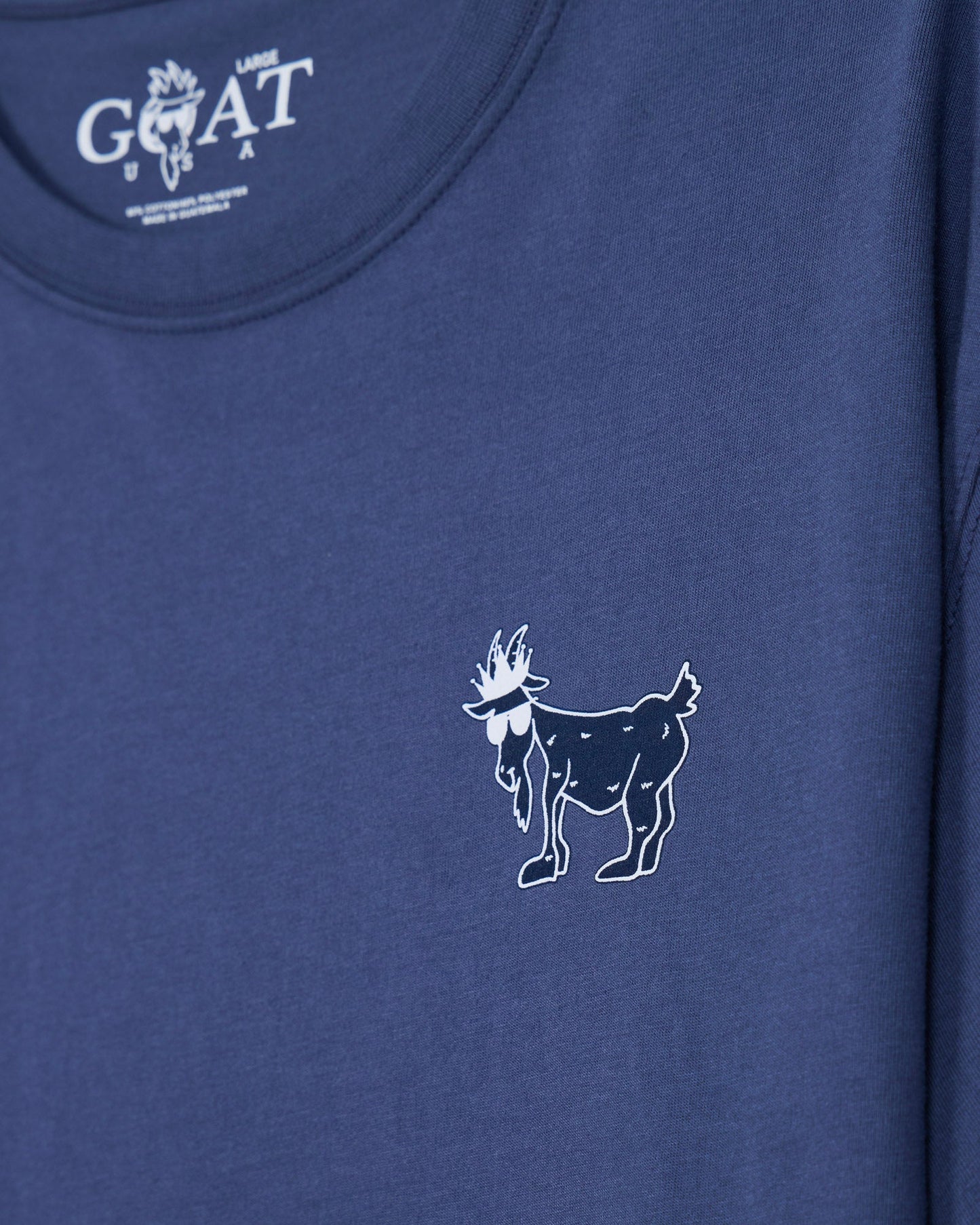 Close-up of a blue t-shirt featuring a small white line-art goat graphic on the chest and a white 'GOAT USA' logo printed inside the collar.#color_navy