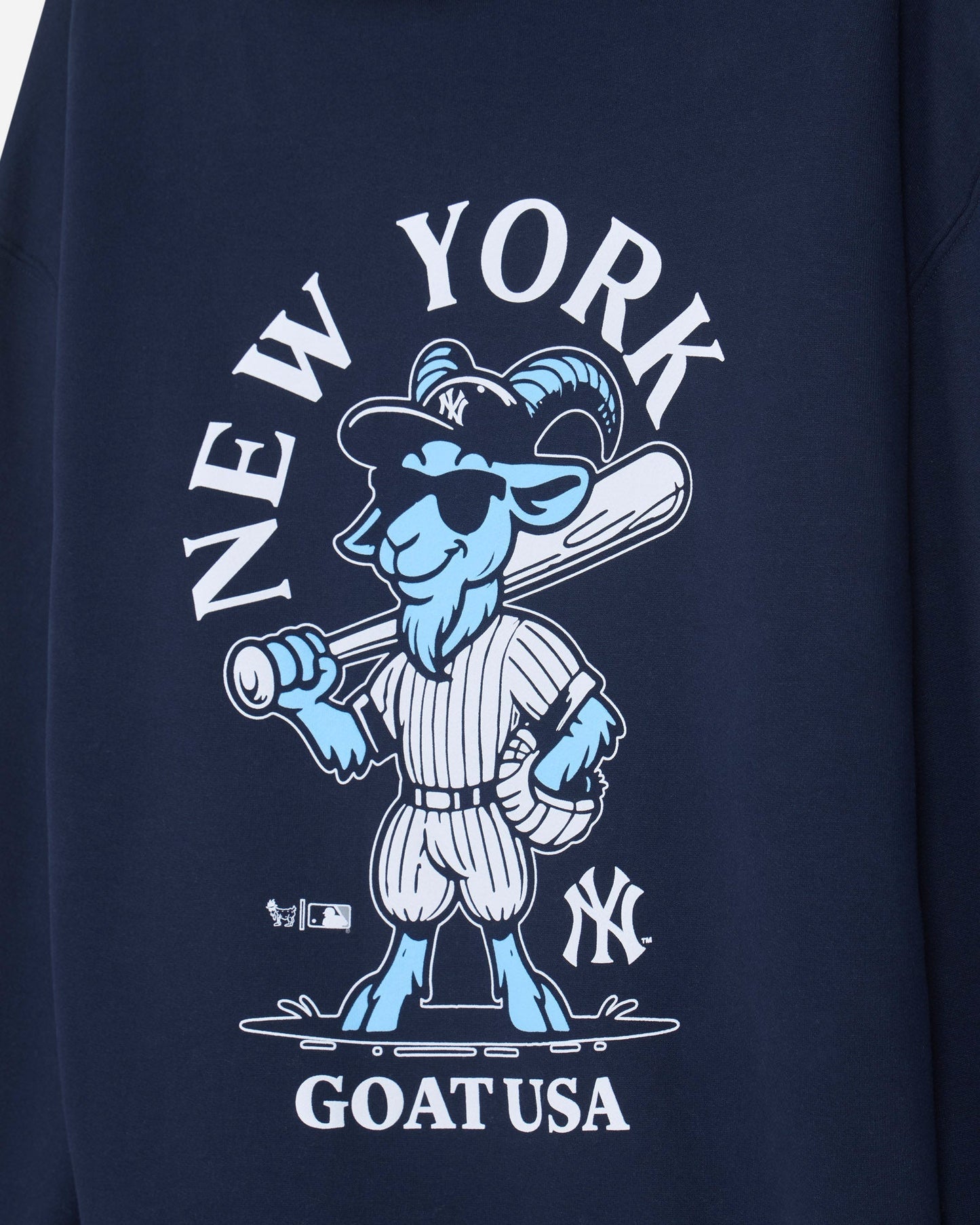 Close-up of a navy blue garment featuring a large front graphic with the words 'NEW YORK' and 'GOAT USA' in white, surrounding a cartoon goat baseball player in blue and white.